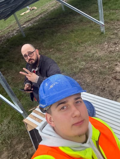This image, taken at Baldurgan Hill, Ireland, depicts two males at a solar panel installation site. In the foreground, a young adult male with fair skin, dark hair, and a slight goatee is shown from the chest up, looking directly at the camera. He wears a dirty blue hard hat, a white hoodie, and an orange and yellow high-visibility vest. Behind him and to his left, an adult male with a bald crown, dark beard, and glasses looks towards the camera, holding a dark smartphone in his right hand and forming a peace sign with his left hand. He wears a dark jacket or shirt over a lighter garment. The men are positioned amidst metal racking structures, likely supports for solar panels, some of which are visible. The ground consists of disturbed earth, mud, and sparse green grass. The overall environment suggests an active outdoor construction or energy development site.