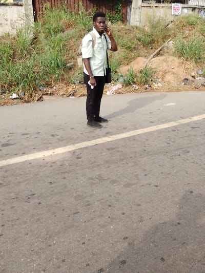 A young adult male stands on an asphalt road in Irete, Nigeria, facing slightly left from the camera's perspective. He is wearing a light green short-sleeved collared shirt, dark trousers, and dark sneakers. A light-colored backpack is worn on his left shoulder, and a dark messenger bag is slung across his right side. He holds a dark smartphone to his right ear, appearing to be on a call, and a small white object in his left hand. The asphalt road features a faded yellow line running horizontally across its surface and shows various dark stains. To the man's right, an overgrown embankment slopes upward, densely covered with green grass, weeds, and exposed reddish-brown soil. The base of the embankment is heavily littered with plastic and other debris. In the background, behind the vegetation, partial views of concrete structures, a reddish-brown metal gate, and a small white sign with red text are visible. The scene is illuminated by strong daylight, casting a distinct shadow of the man onto the road surface.