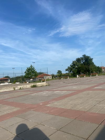 The image displays an outdoor scene in Bryastovets, Bulgaria, characterized by a paved pathway, green parkland, and a clear blue sky. The pathway, made of grey and reddish square paving stones arranged in a diagonal pattern, occupies the right side of the frame and extends into the distance. A dark shadow, likely cast by the photographer, falls across the immediate foreground of the path.

To the left of the pathway, a landscaped area features several mature green trees and multiple wooden park benches with metal frames. Beyond these, a chain-link fence encloses what appears to be a sports court, indicated by two black-framed light fixtures on poles above the fence line. In the background, distant buildings with tiled roofs are partially visible among the foliage.

The sky is a bright, vibrant blue with scattered, wispy white cirrus clouds. A few small, dark specks, possibly birds, are discernible against the sky. The bright daylight and visible shadows suggest the photograph was taken on a sunny late morning or afternoon. No specific human activity is depicted, contributing to a tranquil and open atmosphere. There is no visible text.