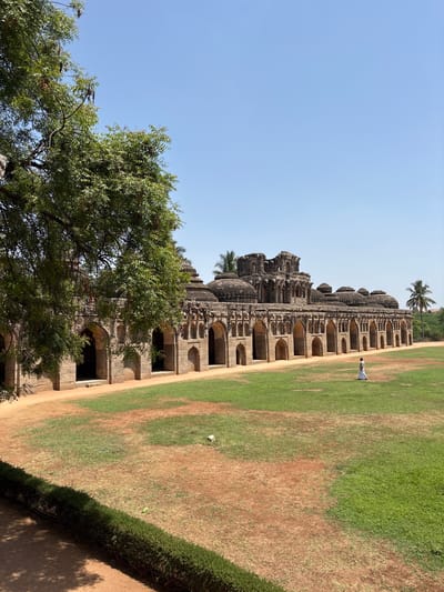 The image shows the Elephant Stables in the city of Kamalapura, India, a long, single-story building with multiple domed chambers. The structure is made of stone with arched openings. A person in white stands near the building. A large grassy area, transitioning from green to brown soil, stretches beside the stables. A tree is in the lower left corner. The sky is a clear blue.