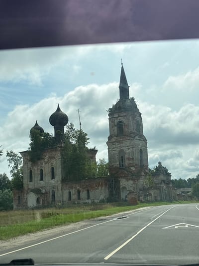 The image shows an abandoned, dilapidated church complex situated in Pryamik, Russia. The architecture features multiple domes and a tall bell tower, all in a state of severe disrepair with crumbling brickwork and vegetation growing over the structures. The church is located adjacent to a paved road, with a grassy verge between the road and the building. The sky is partly cloudy, with cumulus clouds against a light blue background. The scene appears to be captured from a vehicle, as indicated by the darkened upper portion of the image, possibly a glare or tint on a car window. There are no people visible in the image.