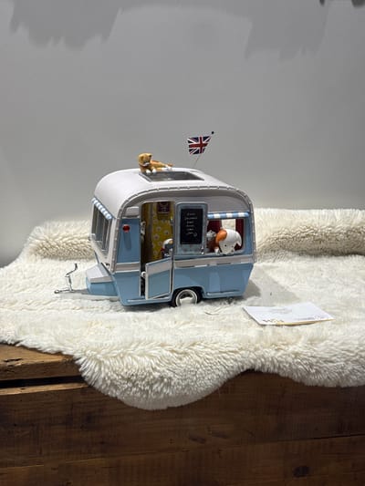 A detailed shot shows a miniature camper van displayed on a white, fluffy rug next to a wooden surface. The camper is predominantly white and blue, decorated with a small Union Jack flag. A cat figurine is positioned hanging out of one of the windows, and a pig figurine is visible through another window. A small chalkboard displaying a menu is affixed to the side of the camper. An envelope sits on the rug next to the camper. While the image does not explicitly show Windermere, United Kingdom, the Union Jack flag suggests a British connection.
