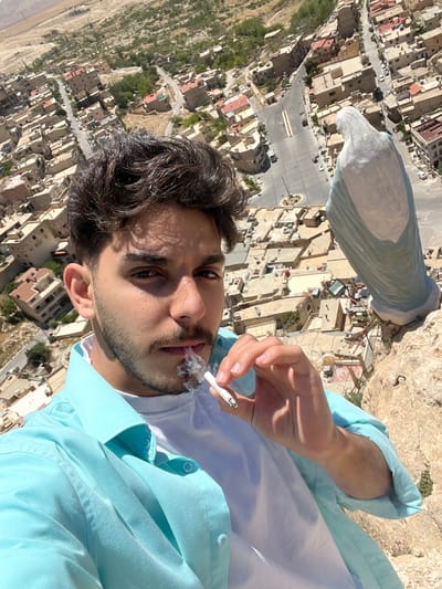 A young man is smoking a cigarette in an outdoor setting, with a cityscape in the background. He is wearing a white t-shirt and a light blue button-down shirt. The cityscape is identifiable as Ma'loula, Syria, given the distinct architectural style and terrain. The photograph is taken from a high-angle perspective, possibly from a cliff or elevated location. The sun is shining.