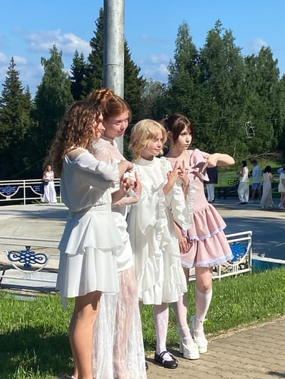 Four young women in light-colored dresses are standing on a paved area with green grass to their left and a line of trees behind them under a bright blue sky. The woman on the far left has curly brown hair and is wearing a white off-the-shoulder dress with ruffled layers. Next to her is a woman with reddish-brown hair and a white lace dress. The third woman from the left has blonde hair and a white ruffled dress. The woman on the far right wears a pink ruffled dress and white socks with white platform shoes. She is pointing her finger to the right. In the background, other people are visible, some standing near a white railing. The environment appears to be an outdoor recreational area or park, possibly part of the town of Galevo, Russia.