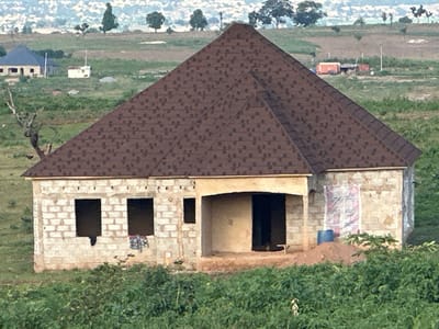 A new house is under construction in a rural setting in Gidan Waziri, Nigeria. The house has a prominent dark brown shingled roof and walls made of concrete blocks. Two large, dark rectangular window openings are visible on the left side of the house. The entrance is an arched opening with a dark door. There is a pile of sand and a blue bucket near the entrance. A white plastic sheet with a pink and white pattern is leaning against the wall on the right side of the house. The surrounding landscape is green and hilly, with some trees and other buildings in the distance. The scene appears to be set in the late afternoon, with soft lighting and long shadows, suggesting a clear day. No people are visible in the image. The overall impression is one of ongoing development and a building in progress.