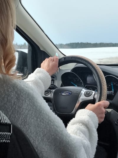 The image depicts the interior of a Ford vehicle, observed from a rear-passenger perspective, situated in Kvarsa, Russia. The driver, visible from behind their left shoulder, has light-colored, shoulder-length hair and is wearing a light-toned, textured jacket. A black and white striped seatbelt is visible across their left shoulder. Both hands are positioned on the vehicle's steering wheel: the left hand, displaying light blue/green iridescent nail polish, is near the 10 o'clock position, and the right hand, wearing a ring on the fourth finger, is near the 3 o'clock position. The steering wheel is black with a woven brown and black cover on its rim and features a central Ford emblem. The instrument cluster shows a tachometer needle slightly above 0 RPM and a speedometer needle indicating approximately 50 units (likely km/h). A central digital display shows "0°C," "D-G," and "180 km." The exterior environment, viewed through the windshield, reveals a snow-covered landscape stretching to a distant line of dark evergreen trees, under an overcast, light grey sky.