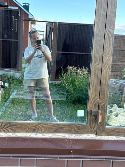 An individual stands in front of a reflective surface, taking a mirror selfie. They are wearing a white t-shirt with green lettering and beige shorts, with their hair pulled up in a bun. The background, visible in the reflection, shows a brick house with a green roof, a dark wooden fence, and green grass. The setting appears to be a residential outdoor area in the village of Pyachanki, Russia. The individual is holding a smartphone in their right hand, and their left hand is by their side. There are no other individuals or significant interactions depicted. The overall environment suggests a casual, possibly domestic, outdoor setting on a clear day.