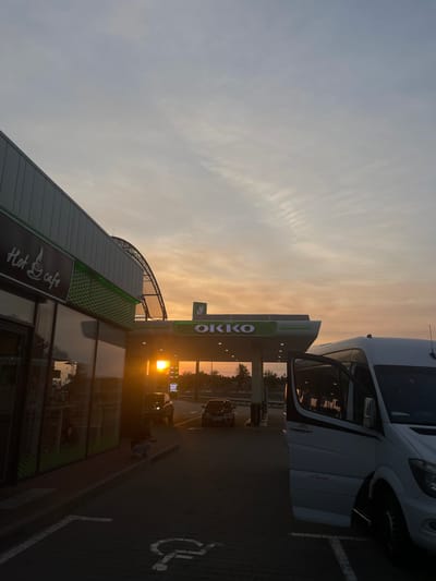 The image depicts a gas station in Shchaslyve, Ukraine, during sunset. A white van with its side door open is parked in the foreground on the right, with a person visible inside. In the middle ground, a gas station canopy with the "OKKO" logo is illuminated by the setting sun. Cars are visible at the pumps. On the left, a building with a "Hot cafe" sign is partially visible. The sky is filled with soft, warm colors of sunset, with wispy clouds. The ground is paved with interlocking bricks, and parking spaces are marked.