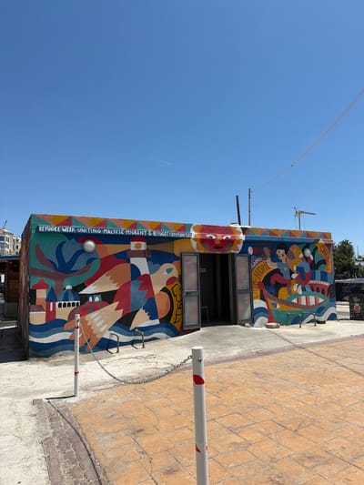 The image depicts a brightly colored building with a mural on its facade, located in Gżira, Malta. The mural, painted in a vibrant abstract style, features elements such as buildings, birds, and people in a boat, symbolizing unity and migration. Text on the mural reads "REFUGEE WEEK UNITING MALTESE, MIGRANT & REFUGEE COMMUNITIES" and "QALBNA OUR HOME". The building appears to be a public restroom, with double doors leading into an open interior. In front of the building, there are bollards with chains and a paved area with orange and grey tiles. The sky above is clear and blue, with a faint airplane trail visible. The overall scene suggests a public space decorated with art to promote social awareness.