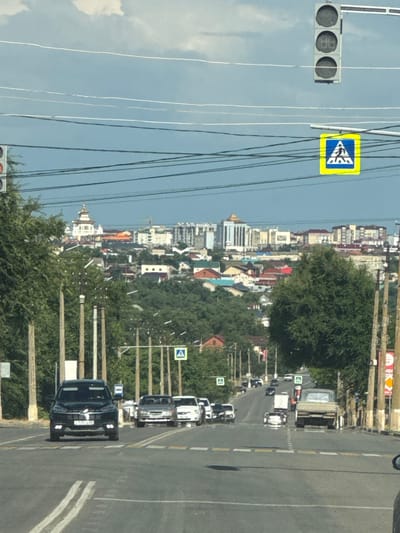 The image shows a street scene in a town or city, likely in Russia, as indicated by the "Дорожный" context. The photograph is taken from a slightly elevated perspective, looking down a road lined with trees and buildings. The road appears to be a two-lane road with a white dashed line dividing the lanes. Several vehicles are visible on the road, including cars and a truck, suggesting that this is a moderately busy street. One black SUV in the foreground has a license plate that reads "T239CA 777". There are also pedestrian crossing signs visible, indicating that this is a safe area for pedestrians to cross.
The buildings lining the street appear to be a mix of residential and commercial structures, with various architectural styles and colors. Some buildings have multi-story designs, while others are smaller, more traditional homes. Lush green trees line the sides of the road and are interspersed with the buildings, adding a natural element to the urban landscape.
In the upper left corner, a traffic light with three circular lights is visible. Next to it, a yellow square sign with a blue triangle and a white symbol is mounted on a pole. Overhead, numerous electrical wires crisscross the sky, which is partially obscured by clouds, suggesting it might be a partly cloudy day.
The overall impression is a typical day in a Russian town or city, with everyday activities like driving and commuting taking place. The lighting suggests it's daytime, possibly late morning or early afternoon, given the bright sunlight and clear shadows. There are no discernible emotions expressed, as the scene is focused on the environment and the objects within it. The image is captured in a vertical orientation.