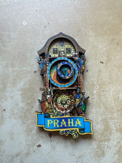 A three-dimensional, multi-layered souvenir magnet, designed to replicate the Prague Astronomical Clock (Orloj), is centrally positioned on a textured, light beige surface. The object is approximately 10-12 cm in height, constructed from a rigid material like wood or plastic, and features printed or painted details.

The top section of the souvenir depicts an arched architectural element with small rectangular panels and a central figure. Below this, the upper astronomical dial (astrolabe) is represented by concentric blue, black, and earth-toned rings, displaying Roman numerals and symbolic representations. Below the astrolabe, the lower calendar plate shows a circular arrangement of twelve pictorial medallions, likely representing months or zodiac signs, surrounding a central crest.

Stylized human figures in colored robes flank both dials: figures in blue and reddish-brown are visible on the left, while figures in blue and green attire are on the right. A prominent blue banner with a yellow border at the base of the souvenir displays "PRAHA" in yellow capital letters, accompanied by decorative scrollwork.

The background surface is a flat, light beige material exhibiting numerous fine scratches, irregular dark speckles, and subtle tonal variations, consistent with a worn countertop or similar surface. The lighting is even, producing no significant shadows. This image captures the described object in Neratovice, Czechia.