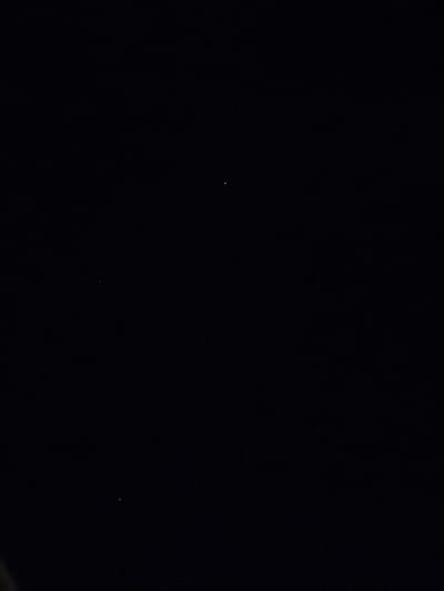 The scene, purportedly located in Villa di Gemmano, Italy, is overwhelmingly dark, presenting as an undifferentiated black field across the majority of the frame. Against this dark environment, three minute points of light are visible. A bright white point is positioned in the upper-central portion of the image. A second, dimmer, bluish point of light is situated in the lower-left quadrant. A third, very faint white point is discernible in the upper-right quadrant. A subtle, dark, out-of-focus curved element is present at the extreme bottom-left edge of the frame. No people, specific subjects beyond the light sources, or any actions and interactions are discernible.