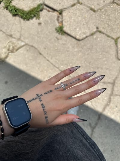 A person's hand and wrist are positioned against a background of hexagonal stone pavement and a dark denim garment. The hand features long, almond-shaped nails with a clear base and black tips, each adorned with a small black dot. A silver ring is on the ring finger. There are tattoos on the hand and wrist, including the words "LOVE AFTER PAIN HERE" and "RAIN SUN" with the word "AFTER" written on the wrist. A black smartwatch is on the wrist, displaying the time "13:40" and the date "MAY 26 TUE". A beaded bracelet is visible below the watch. The photo is taken outdoors in daylight. There is no indication in the image that determines the location is in the city of Mednikarovo, Bulgaria.