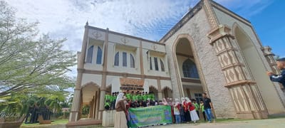 In the city of Kruengmate, Indonesia, a group of children and adults stand outside a cream-colored building with a large arched entrance, possibly a museum. Many of the children are wearing bright green shirts and headscarves, suggesting they are on a school trip. Some adults are also present, including a woman in a light-colored hijab and a man in a black shirt and jeans. In front of the group, a large green banner with text in Indonesian reads "STUDY TOUR & WISATA EDUKASI SEJARAH MIN 10 ACEH UTARA," indicating a historical education study tour from MIN 10 Aceh Utara. The building's architecture features arched windows and decorative stone accents. To the left of the building, a large tree with sparse leaves casts a shadow, and in the background, there are palm trees and other greenery. The sky is partly cloudy, with white clouds against a blue background.