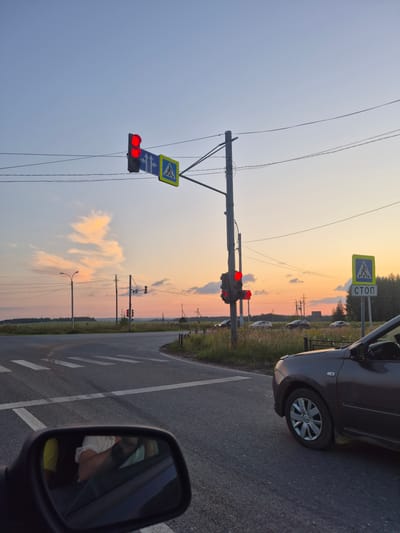The image shows a road intersection at dusk in Novoe Mart'yanovo, Russia. A traffic light displays a red signal, and a pedestrian crossing is marked on the asphalt. A dark-colored car is visible on the right side of the frame, and the reflection of a person wearing a white shirt and yellow garment is seen in the car's side mirror. In the background, there are power lines, utility poles, and a treeline. The sky is a gradient of blue and orange, with some wispy clouds. The general atmosphere is that of a rural or suburban road.