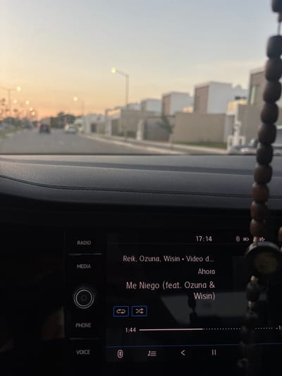 A car dashboard features a music player displaying the song "Me Niego" by Reik, Ozuna, and Wisin. The time is 17:14, and the music is playing at 1:44. Options for Radio, Media, Phone, and Voice are visible. Through the window, a street with houses and streetlights is visible in a light-filled environment. A strand of beads hangs in the foreground.