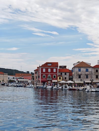 A view of the Stari Grad waterfront in Croatia shows a calm blue body of water in the foreground, reflecting the sky and buildings. In the background, a row of buildings lines the shore. The buildings vary in architectural style, with some featuring red tile roofs and pastel-colored facades, while others are constructed with stone. Several boats are docked along the waterfront, ranging from small motorboats to larger vessels. People can be seen walking along the promenade and sitting at outdoor cafes. The sky above is a mix of blue and white clouds. The overall scene conveys a peaceful, Mediterranean atmosphere.