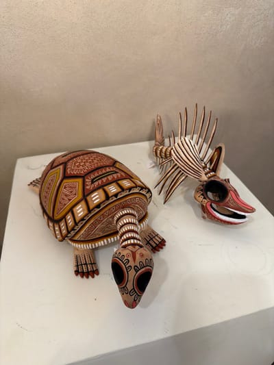 The image displays two intricately carved and painted wooden figures, likely alebrijes, against a textured, light-colored wall, possibly in a corner or an alcove. The scene is static, showing the figures on display.
The top figure resembles a fantastical creature with an elongated body, spiky projections along its back, and an open mouth with visible teeth. It is predominantly painted in shades of beige, brown, and white, with red accents around its mouth and eyes, and features delicate striped and dotted patterns.
Below it is a larger figure of a turtle. Its shell is adorned with elaborate geometric patterns in shades of brown, yellow, and white, including triangles, squares, and dotted lines. The turtle's head, limbs, and tail also feature similar decorative motifs and colors, with black for its eyes. Both figures exhibit a characteristic style of Mexican folk art known for vibrant colors and detailed designs.
No human subjects are present, and no specific activity is taking place beyond the display of the art pieces. There is no visible text in the image. The setting appears to be indoors, potentially a shop or gallery, consistent with the given location context of San Martín Tilcajete, Mexico, a town famous for its alebrije carvings.