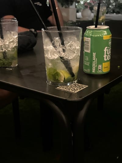 In Vera Playa, Spain, a black, reflective table holds two drinks and a green soda can. A tall, clear glass on the left is filled with ice and what appears to be a mojito, with mint leaves and lime visible. A second, similar glass on the right contains ice and lime slices, also appearing to be a mojito. A green soda can with the text "fuze tea" and "RECICLAME Soy reciclable" is positioned to the right of the second glass, with "Limon" written below a yellow rectangle. A black QR code sticker is affixed to the table in front of the second glass. The background is dark and out of focus, suggesting an evening setting.