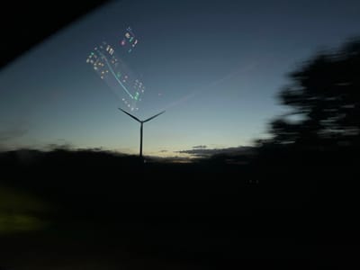 The image depicts a twilight or early evening scene located in Châtenay, France, captured from inside a moving vehicle. A three-bladed wind turbine, dark in silhouette, stands centrally against the sky, its blades oriented roughly diagonally. The sky transitions from a deep blue at the top to a lighter blue-green hue near the horizon, with faint orange and yellow light visible at the lowest point of the sky and scattered dark clouds horizontally. The foreground and midground consist of a very dark, indistinct landscape, suggesting fields or low-lying terrain, rendering specific details unclear. On the far right, a vertical mass of dark, motion-blurred foliage, likely trees, indicates rapid movement relative to the camera. Overlaying the upper-left and central sky is a distinct reflection of a vehicle's internal display screen, showing what appears to be a navigation interface with illuminated green lines, yellow and orange points, and white alphanumeric text, typical of a global positioning system. No people are visible.
