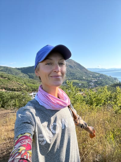 A woman stands on a grassy hillside with a panoramic view of a coastal city and mountains. She is wearing a blue baseball cap, a gray sweatshirt with white lettering, and a pink patterned neck gaiter. Her left arm is covered by a tattoo sleeve with flame and heart designs. She is smiling and holding a small orange object in her right hand. The background shows a densely populated hillside with buildings, greenery, and the sea. The sky is clear and blue. The scene is set in Lazurnoe, Ukraine.