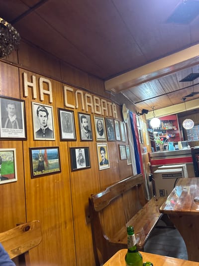 The image depicts the interior of a commercial establishment, likely a restaurant or tavern, situated in the city of Sushitsa, Bulgaria. The walls and ceiling are covered with light-toned varnished wood paneling. A prominent display on the left wall features a collection of framed portraits, primarily historical black and white photographs of men, alongside a few color paintings, including one with Cyrillic text identifying "Райна Попгеоргиева." Above these portraits, large, raised light-colored Cyrillic letters spell "НА СЛАВАТА" (TO THE GLORY). A vertically oriented Bulgarian flag (white, green, red stripes) hangs on the wall to the right of the portraits. Varnished wooden benches with backrests are positioned along the wall, accompanied by matching wooden tables. A green glass bottle rests on a table in the foreground right. In the background right, a bar area is visible, featuring a counter with red and silver accents, shelves stocked with bottles and glassware, a potted plant containing red flowers, and various light fixtures. A cardboard box labeled "PROFESSIONAL SOUND SYSTEM" is located on the floor near the bar. A partial view of a person's limb is visible in the far left foreground.