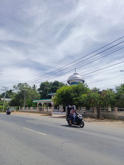 This is a wide shot of a paved road in Blangkubu, Indonesia. A modern mosque with a large, silver, domed roof and green accents is visible on the right side of the road, behind a fence and surrounded by lush green trees. Two motorcycles are on the road. The motorcycle in the foreground carries three people: a driver in a helmet, a passenger behind them, and a child seated in front of the driver. A second motorcycle with a single rider is further down the road on the left. Overhead, multiple electrical wires stretch across the frame against a sky with wispy white clouds. The overall impression is a sunny day in a semi-rural area.