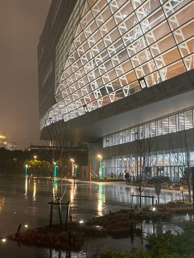 The image captures a modern urban scene at night in Yanqiao, China, under an overcast sky, with surfaces appearing wet, indicating recent or ongoing precipitation. A large, contemporary building dominates the right and upper frame. Its upper levels feature a distinctive glass facade with a white geometric grid pattern, appearing as a series of interlocking triangles and rectangles, illuminated from within by warm light. One window on this upper section is partially open.
The building's lower level includes a prominent overhang and a ground floor entirely composed of large glass windows. The interior of this ground floor is brightly lit, revealing an open space where approximately six to eight individuals are gathered, their actions indistinct.
The ground area in front of the building is a wide, wet paved plaza or walkway, highly reflective of the surrounding lights. Several young, bare-branched trees, some with support structures, are interspersed within the pavement and in small, dark landscaped islands of low-lying vegetation. Numerous streetlights emit yellow and green light, casting long, vibrant reflections across the wet surface.
Two individuals are visible outdoors on the paved area: one in the midground walks left, potentially holding an umbrella, while another is further back near a line of green streetlights. A reddish-brown vertical panel is affixed to the building's exterior wall on the ground level, left of the main entrance area. In the far background on the left, a portion of another illuminated building with a golden glow is visible.