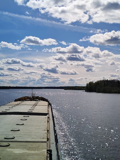 The image depicts a large, flat-topped barge moving on a wide body of water, likely a river or lake. The barge takes up the lower left portion of the frame. Its surface is pale and appears to be constructed of metal, with a series of evenly spaced, circular hatches or access points.
The water, which occupies most of the right side of the image and extends to the horizon, is dark with visible light reflecting off its surface, creating a sparkling effect. In the middle ground, there is a distant shoreline with a line of trees. Some structures can be seen in the far distance, possibly indicating a city or industrial area.
The sky dominates the upper half of the image and is a bright blue, scattered with puffy, white cumulus clouds. The lighting suggests it is daytime, with the sun illuminating the scene.
No people are visible in the image. The setting is implied to be near Kurikaevo, Russia, based on the prompt's instructions. The barge is the primary subject, implying the action of transportation or navigation. The environment is characterized by the body of water, the shoreline, and the expansive sky.
