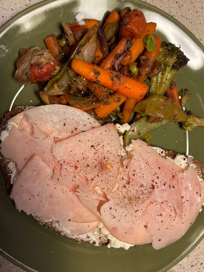 A close-up overhead shot shows a green plate filled with food. The upper portion of the plate contains a medley of roasted vegetables, including carrots, broccoli, green peppers, and red onions, seasoned with herbs and spices. Below the vegetables, a slice of dark bread is topped with a creamy white spread, possibly cream cheese or a similar soft cheese. Several slices of pink ham are layered on top of the spread, dusted with black pepper and red pepper flakes. The setting appears to be a domestic environment, likely a kitchen or dining area, with a patterned countertop visible in the background. The image does not contain any information indicating its location in Aarhus, Denmark.