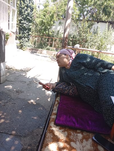 A woman wearing a pink headscarf and a dark green patterned dress is lying on a patterned mat outdoors. She is holding a red mobile phone in her right hand and appears to be looking at it. The woman is resting on a purple cushion. The background shows lush green foliage and trees, a white wall, and a metal railing. Sunlight casts shadows on the asphalt ground. The setting appears to be a residential outdoor area in the city of Hisor, Tajikistan, possibly a courtyard.