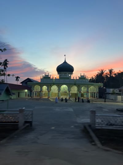 The image depicts a mosque in Matangpanyang, Indonesia, during sunset. The sky displays a gradient of blue, pink, and orange hues. The mosque features a prominent dark green dome topped with a crescent moon and star symbol. The building has a white facade with green accents and is supported by arches. Several people, some wearing light-colored clothing, are visible in front of the mosque, along with a few motorcycles. Palm trees and other vegetation surround the mosque, and the ground in front appears to be paved. The scene is illuminated by the twilight sky and some artificial lighting from the mosque.