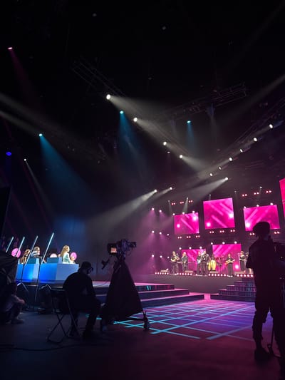 The image depicts a wide shot of a stage set up for a performance or broadcast event, potentially at the "Domodedovo Airport" in Nemchinovka, Russia. The stage is illuminated with vibrant pink and blue lighting, with spotlights cutting through the dark atmosphere from above. In the foreground, a person is seated on a folding chair, facing away from the camera, appearing to be part of the production crew. To the right, a large professional camera on a tripod is positioned, also facing the stage.

The stage itself features several tiered platforms with reflective surfaces, some of which are lit with a grid pattern of blue and pink light. Behind the platforms, a band of several musicians is performing, playing instruments like guitars and drums. Large LED screens displaying abstract pink graphics are mounted behind the performers. To the left of the stage, a group of individuals are seated behind a long blue counter, seemingly as judges or contestants. The overall environment suggests a well-equipped studio or performance venue.