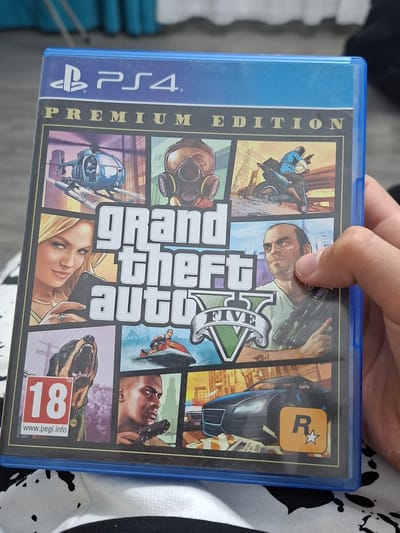 The image shows the cover art of the video game Grand Theft Auto V: Premium Edition for PlayStation 4. The cover features a collage of scenes and characters from the game, including a helicopter, a person in a gas mask, a person on a motorcycle, a woman holding a phone, a snarling dog with a PEGI 18 rating, a person on a jet ski, a man holding a rifle, and a sports car. The game's title, "Grand Theft Auto V," is prominently displayed in the center. The PS4 logo and "PREMIUM EDITION" are visible at the top. The game case is held by a person's hand, with fingers visible on the right side of the image. The background is blurred but appears to be an indoor setting with patterned fabric visible at the bottom. There is no information in the image or its context that places this scene in Biryusinsk, Russia.