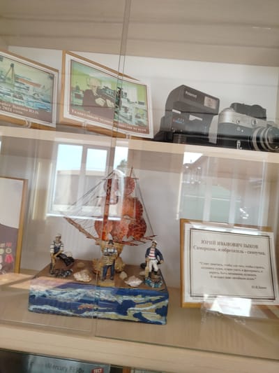 This is a medium-shot, eye-level photograph of a display case containing historical artifacts in Yashkino, Russia. The display case is made of wood and glass. On the top shelf, a Polaroid camera and a silver and black camera are visible. Below this, a diorama of a ship with two figurines is displayed on a blue and white base. To the right of the diorama, a framed placard with text in Russian, bearing the name "ЮРИЙ ИВАНОВИЧ ЗЫКОВ," is displayed. The placard contains a quote and his signature. The background of the display case shows a window with reflections of buildings outside. The bottom shelf of the display case appears to contain books.