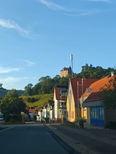 The image depicts a street scene in Schieder-Schwalenberg, Germany, likely during late afternoon due to the warm, low-angled sunlight. A paved road occupies the foreground, curving slightly to the left. On the right side of the road, a row of buildings with red tile roofs lines the street. These buildings exhibit a mix of architectural styles, with some featuring yellow brick facades and others white exteriors. One building has a prominent blue-windowed section. Behind the buildings, a steep, green hillside covered in trees rises, with a castle-like structure and a tall cell tower visible on its summit. On the left side of the road, more buildings are visible, set back from the road and partially obscured by trees. A few cars are parked along the street or driveways. The sky is a clear, bright blue with a few wispy clouds. The overall impression is of a quiet, residential area in a hilly landscape.