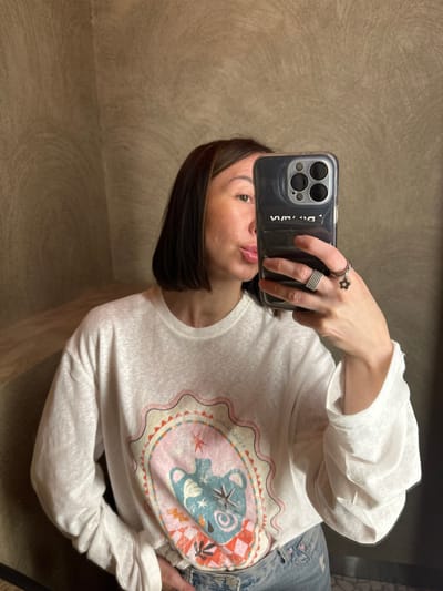 This is a mirror selfie taken indoors in the city of Усады, Russia. A woman with short, dark brown hair and lighter highlights is depicted from the chest up, holding a dark-colored smartphone with a clear case in her right hand. The phone partially obscures the right side of her face. She is looking towards the camera's reflection, with her left eye visible and her lips slightly parted, her tongue partially extended.
She wears a loose-fitting, white, long-sleeve t-shirt with a colorful, intricate graphic print centered on the chest. The graphic features an oval frame containing abstract shapes and a stylized animal figure in shades of pink, blue, orange, and red. Her lower body is partially visible, showing blue denim jeans. On her right ring finger, she wears a multi-banded silver-colored ring, and on her right pinky finger, she wears a silver-colored ring with a dangling, dark flower charm.
The background consists of textured walls in a light brownish-beige or taupe color, displaying distinct swirling patterns. A corner where two walls meet is visible on the left. A small portion of a dark, possibly tiled, floor is visible at the bottom right. The smartphone's triple camera lenses are visible on the back, and inverted text is discernible on the phone case due to the reflection.