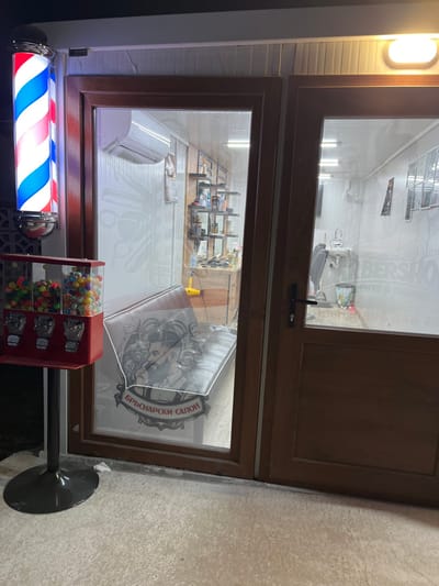 This image captures the exterior of a barbershop at night in the city of Branichevo, Bulgaria. The illuminated storefront features a vertical, rotating red, white, and blue barber pole on the left, adjacent to a red stand-alone gumball machine filled with multi-colored spherical candies. The entrance consists of a brown-framed glass panel on the left and a matching opaque wooden door on the right.
Through the glass, the interior of the brightly lit barbershop is visible. A large white decal depicting a bearded man and the Cyrillic text "БРЪСНАРСКИ САЛОН" (Barbershop) is affixed to the inside of the left glass panel. Behind this, a dark bench or sofa is present. Further inside, an air conditioning unit is mounted high on the wall, above shelving units stocked with grooming products and a workstation with a yellow hairdryer. On the right side of the interior, a barber chair is positioned in front of a white wall-mounted sink, with multiple framed items on the far right wall. Partial English text, including "BARBERSHOP," is also visible on the interior side of the right glass panel. The ceiling is equipped with strip lighting, illuminating the light-colored floor. The exterior ground directly in front of the shop is a concrete surface. No individuals are present in the scene.