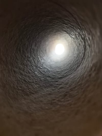 The image depicts a close-up, low-angle shot looking down the interior of a cylindrical object, possibly a cardboard tube. The walls of the tube are textured and appear to be a dark, muted brown color, with visible striations and variations in shading. Light is emanating from the far end of the tube, creating a bright, circular focal point that diminishes in intensity as it recedes into the darkness. The overall impression is one of depth and perspective, drawing the viewer's eye towards the distant light source. There are no people or specific landmarks visible in this image. The scene is entirely abstract and focuses on the texture and form of the interior of the tube and the light at its end.