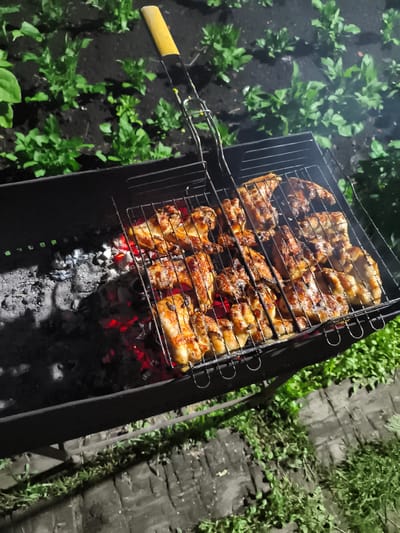 An overhead, medium shot captures a close-up of chicken pieces cooking on a grill over glowing coals. The chicken, seasoned and browned, rests on a black metal grill basket, which is positioned above a dark, metal barbecue grill. The coals beneath glow a vibrant red, casting a warm light on the food and the grill. To the left of the grill, ashes and embers are visible. The background consists of dark soil with green plants, suggesting an outdoor setting at night. A yellow and black handle of a grilling tool is visible at the top of the frame. The scene appears to be in Il'mino, Russia, a location known for its natural beauty and outdoor recreational activities.