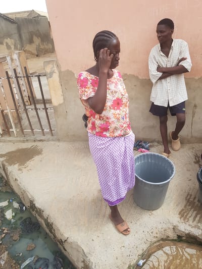 The image shows two people in an outdoor setting, possibly in Dutsinma A, Nigeria. A man wearing a checked shirt and dark shorts lies on a concrete surface with his arms crossed. Next to him, a woman wearing a floral top and a patterned purple skirt stands with her hand near her face. Near the woman is a gray bucket. The location appears to be near a building with a barred window. A waterway filled with trash is visible in the lower right corner.
