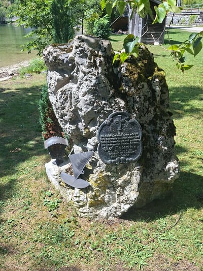 The image shows a monument erected on the shore of a lake, likely in Obertraun, Austria, given the context. The monument consists of a large, rough-hewn rock covered in moss. Attached to the rock is a dark-colored plaque with an inscription and a cross at the top. To the left of the plaque, there is a small evergreen plant, and below it are two metal objects: a lamp-like structure with intricate detailing and a triangular, anchor-like object. In the background, a body of water is visible, with a shoreline of pebbles and grass. Further back, trees and a small wooden structure, possibly a shed, can be seen. The ground around the monument is covered in grass and scattered leaves, with sunlight casting shadows. The overall scene depicts a solemn memorial situated in a natural, lakeside environment.