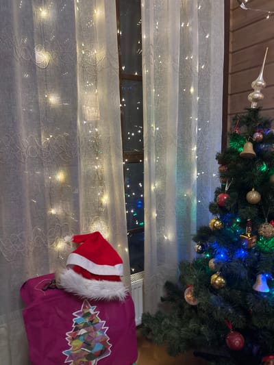 A Christmas tree decorated with lights and ornaments sits above a window covered with sheer, light-strung curtains. A red Santa hat and a magenta sack with colorful geometric designs rest below the window on the floor. The scene evokes a cozy, festive atmosphere, possibly a glimpse into a home celebrating the holiday season. The window reveals a view of distant lights, suggesting it's nighttime.