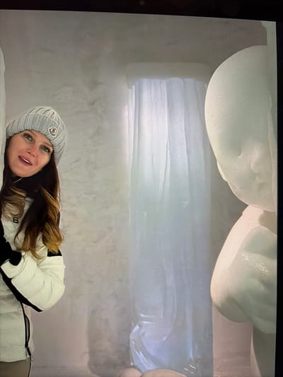 The image captures an indoor scene within an ice structure, characteristic of an ice hotel or art exhibition located in the Kiruna region of Sweden. A female subject is positioned on the left, partially framed by an ice wall, looking directly towards the viewer with a slight smile and parted lips. She has long brown hair and wears a light grey ribbed knit beanie adorned with a circular Moncler logo patch, a black high-collared garment, and a white puffer jacket featuring black accents on the shoulders and sleeves. To the right, a large, translucent ice sculpture depicts the upper half of a stylized human figure, resembling an infant or child, with smooth, rounded features including a head, face, and torso. The background consists of textured ice walls and a prominent vertical formation of translucent ice blocks, creating a curtain-like effect that extends from the ceiling to the floor. The overall environment is illuminated with soft, ambient light, highlighting the crystalline nature of the ice.