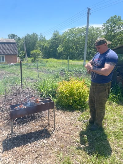 In the city of Yasnoe, Russia, a man wearing a camouflage cap, a blue t-shirt with a graphic print, and camouflage pants stands in a yard preparing to grill. He is holding a metal grill rack, and his arms are covered in tattoos. In the foreground, a rusty barbecue grill is set up with a fire burning inside, and several sticks with food items are placed over the flames. To the left of the grill, a pile of dry branches is stacked. The background shows a green lawn, a fence, a building with a corrugated roof, and lush trees under a clear blue sky. The ground is covered with pebbles and dry grass.