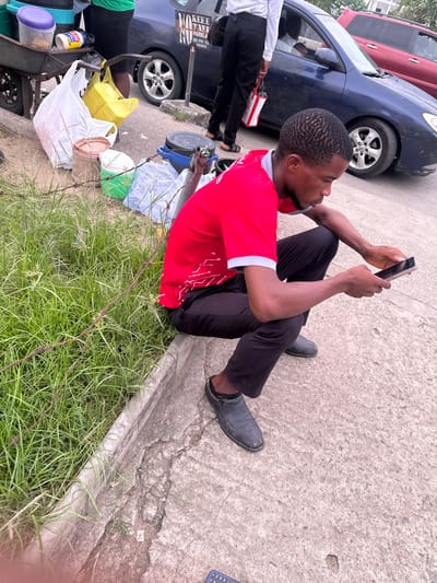 A man in a red shirt and black pants sits on a concrete surface, looking at a cell phone. He wears black shoes. Beside him is a grassy patch. Behind him is a blue car, part of a red vehicle, and a person in a white shirt and black pants. A cart filled with containers, bags, and other items is also visible.