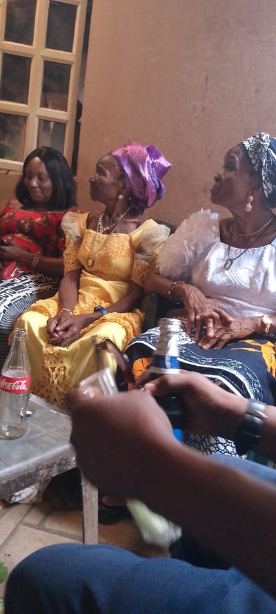The image depicts an interior or sheltered patio scene in Ohafia, Nigeria. An unadorned light brown wall forms the background, with a white-framed, multi-paned window positioned on the upper left. Three women are seated in a row, facing generally towards the right. From left to right: The first woman has dark hair, wears a red patterned top and a black-and-white patterned fabric, and is observed looking downwards while holding a mobile phone in her left hand. The second woman, elderly, wears a yellow dress with sheer sleeves and lace detailing, a purple headwrap (gele), multiple necklaces including a silver choker and a long pendant, earrings, and a blue smartwatch on her left wrist; her hands are clasped. The third woman, also elderly, wears a white ruffled top, a dark blue patterned fabric, a black-and-white patterned headwrap, a necklace with a rectangular pendant, and gold-colored earrings and watch; her hands are clasped. Both elderly women look upwards and to the right. In the immediate foreground, an individual wearing dark blue trousers is partially visible, with hands actively engaged in opening a dark-colored bottle with a blue label, using a bottle-opening tool. An empty glass Coca-Cola bottle rests on a mottled grey utility table in the lower left quadrant of the frame. The floor, partially visible beneath the table, appears tiled. The scene suggests a social gathering or shared activity.