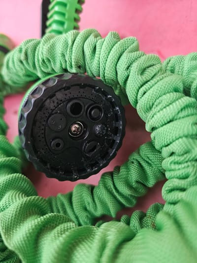 This is a close-up, top-down shot of a coiled green expandable garden hose with a black spray nozzle. The hose is made of a textured, woven material and is coiled in a circular pattern around the nozzle. The nozzle itself has multiple spray settings, indicated by different-sized holes and a central screw. The background is a bright pink surface, likely a table or floor. The lighting appears to be artificial, possibly from an overhead light, casting soft shadows. There are no people or animals visible, and no context for a specific event or time of day is provided. The image focuses solely on the texture and form of the garden hose and nozzle. There is no visible text in the image. The location context of Mäntsälä, Finland, is provided externally and cannot be inferred from the image itself.