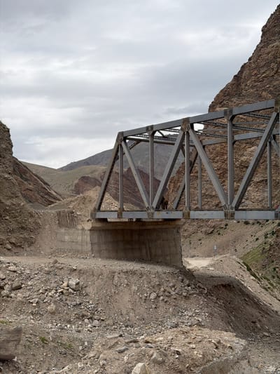 The image depicts a steel truss bridge spanning a rocky, mountainous terrain. The bridge is gray and constructed with a lattice of diagonal beams. It crosses over a dry riverbed or ravine. Concrete abutments support the bridge. The surrounding landscape is characterized by steep, barren slopes covered in loose rocks and gravel. Vegetation is sparse, with a few patches of dry grass and scrub. The sky is overcast with gray clouds. A dirt road winds its way up the mountainside on the right side of the image. There is a single person, appearing to be a male, standing on the bridge, looking away from the camera. Another person is partially visible on the far right of the bridge structure. The scene appears to be in a remote, undeveloped area. The location is identified as Reru, India.