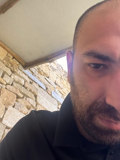 The media file is a close-up, low-angle selfie of a person's face, partially obscured by the camera's proximity and the subject's downward gaze. The person appears to be male, with short-cropped hair and a light beard. One eye is clearly visible, dark and focused, suggesting an intense or contemplative expression. The background is a textured stone wall, likely part of an outdoor structure, with some wooden beams visible on the left. The lighting suggests it might be daytime, possibly outdoors given the natural stone and visible light. The orientation of the image, with the subject's head dominating the frame and looking downwards, creates an unusual perspective. No text is visible in the image. Based on the location context provided, this scene could be in Pliska, Bulgaria, a historical site known for its ancient ruins.